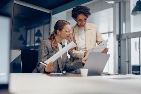 Dekoratives Bild: 2 lächelnde Frauen in einem Büro mit Laptop und Unterlagen